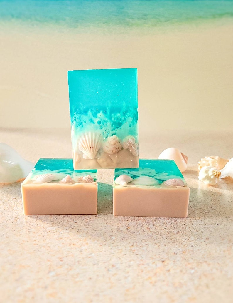 K&ouml;nnte beinhalten: Drei Seifenst&uuml;cke mit Strandmotiv. Die obere Seife ist blau und gr&uuml;n mit wei&szlig;en Muscheln, die in die Seife eingebettet sind. Die beiden unteren Seifen sind beige mit wei&szlig;en Muscheln, die in die Seife eingebettet sind.