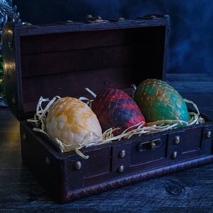 May include: A dark wooden treasure chest with three dragon eggs nestled inside. The eggs are yellow, red, and green, with textured surfaces. The chest is lined with dark red fabric and filled with straw. Decorative metal accents adorn the chest.