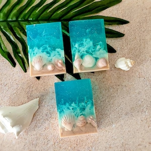 K&ouml;nnte beinhalten: Drei rechteckige Seifenst&uuml;cke mit einem blauen und wei&szlig;en Wirbelmuster und Muscheln, die in die Seife eingebettet sind. Die Seife liegt auf einer hellbeigen Oberfl&auml;che.