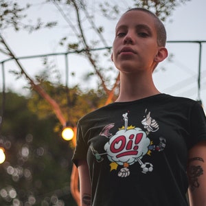 以下が含まれることがあります： 短く刈り込まれた髪をした人が、赤い吹き出しの中に「Oi!」と書かれた漫画のキャラクターの白いグラフィックプリントが施された黒いTシャツを着ています。その人は緑の葉の茂った茂みの前に立っています。