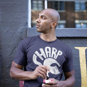 Könnte beinhalten: Ein Mann trägt ein dunkelgraues T-Shirt mit einer weißen Grafik des Wortes "SHARP" im Retro-Stil. Er hält eine Zigarette und eine Schachtel Zigaretten in der Hand.