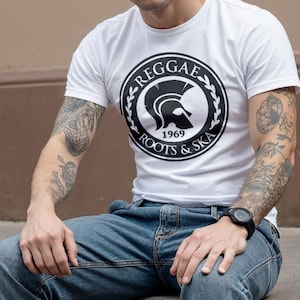 Può includere: Maglietta bianca con grafica nera "Reggae Roots & Ska 1969". La maglietta è abbinata a jeans blu e stivali neri stringati. La persona è seduta su un bordo di cemento.