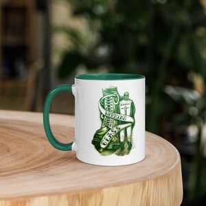 Puede incluir: Taza de cerámica blanca con un borde verde. La taza presenta una ilustración verde de una bota de trabajo con el texto "Working Class" envuelto alrededor. Una persona con uniforme de trabajo se encuentra junto a la bota.