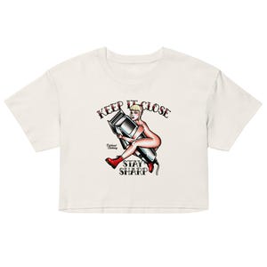 May include: Cream-colored crop top featuring a tattoo-style graphic. The design includes a woman holding a hair clipper, with the words "KEEP IT CLOSE" and "STAY SHARP" in a banner. The woman is wearing red boots.