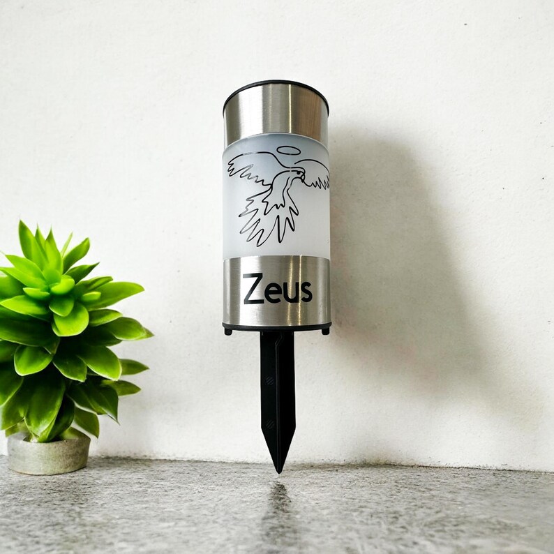 May include: A silver solar powered garden light with a black stake. The light features a frosted glass cylinder with a black and white graphic of a bird with a halo above it. The word "Zeus" is printed on the cylinder.