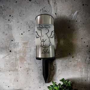 May include: A silver solar-powered memorial light with a white frosted glass insert. The insert features a black line drawing of a rabbit with angel wings and a halo. The light has a black metal stake and the name "Lola" is printed on the bottom.