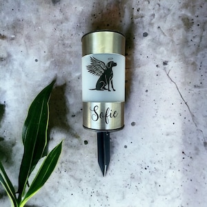 May include: A silver metal memorial solar light with a black silhouette of a dog with wings and a halo. The light has the name "Sofie" engraved on it.
