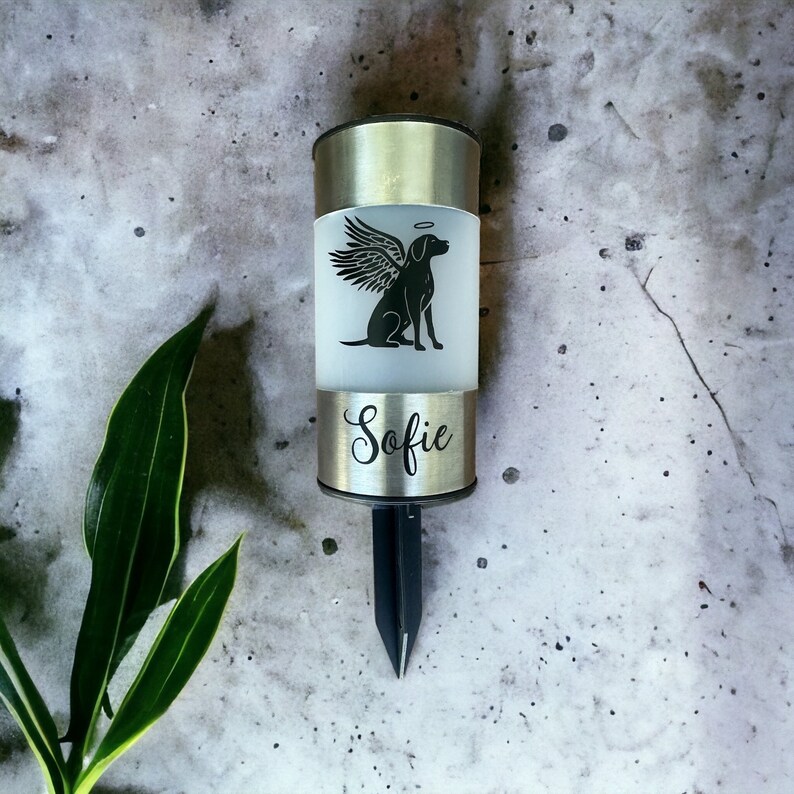 May include: A silver metal memorial solar light with a black silhouette of a dog with wings and a halo. The light has the name "Sofie" engraved on it.