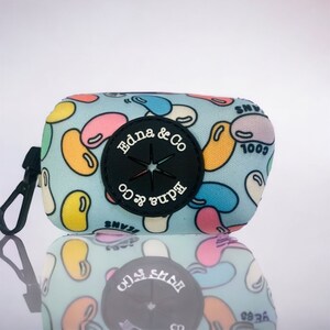 Puede incluir: Un dispensador de bolsas para caca azul y blanco con un patr&oacute;n de gominolas. El dispensador tiene un c&iacute;rculo negro con una estrella y el texto "Edna & Co" en &eacute;l.