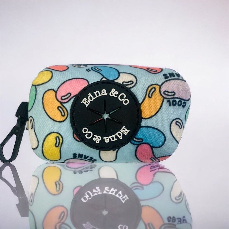 Puede incluir: Un dispensador de bolsas para caca azul y blanco con un patr&oacute;n de gominolas. El dispensador tiene un c&iacute;rculo negro con una estrella y el texto "Edna & Co" en &eacute;l.