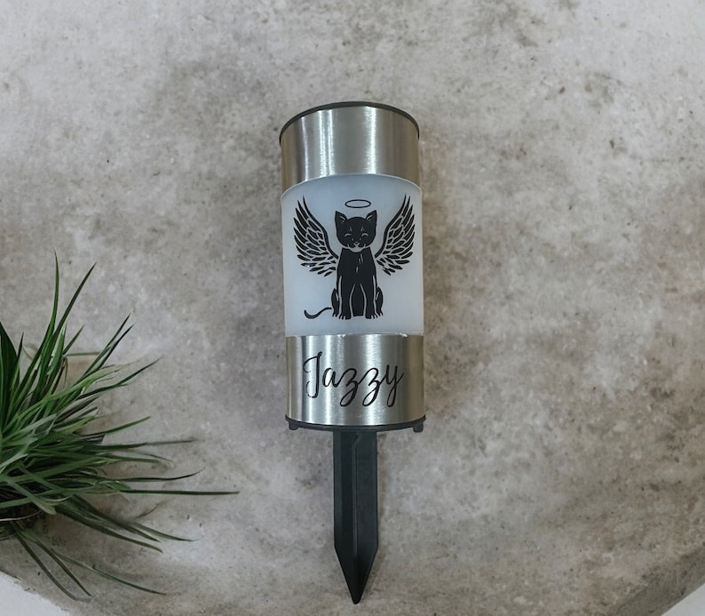 May include: A silver solar-powered memorial light with a black cat with wings and a halo. The light has the name "Jazzy" engraved on it.