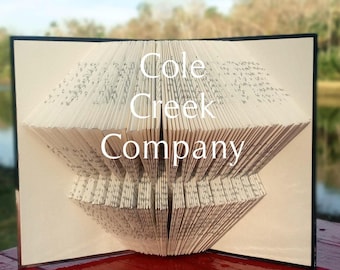 Może przedstawiać: Książka złożona w trójwymiarowy kształt, tworząc wzór z napisem "Cole Creek Company". Książka leży na czerwonej drewnianej powierzchni z rozmytym tłem jeziora i drzew.