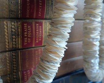 May include: A close-up of a stack of antique books with gold lettering and burgundy spines. A garland made of layered, curled paper is in the foreground. The paper garland is white and has a textured appearance.