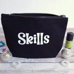 Könnte beinhalten: Ein schwarzes Reißverschlusstäschchen aus Segeltuch mit dem in weißen Buchstaben aufgedruckten Wort Skills. Das Täschchen ist umgeben von zwei kleinen silbernen, stacheligen Sensorikbällen, zwei kleinen Haargummis, zwei Bonbonverpackungen und einem Fläschchen.