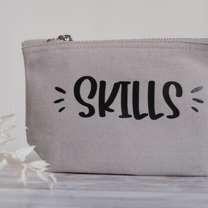 Könnte beinhalten: Eine graue Canvas-Kosmetiktasche mit dem Wort "SKILLS" in Schwarz bedruckt.