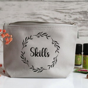 Könnte beinhalten: Eine hellgraue Reißverschlusstasche mit dem Wort "Skills" in schwarzer Schrift, umgeben von einem schwarzen Blumenkranz. Mehrere ätherische Ölflaschen und andere kleine Gegenstände sind in der Nähe angeordnet.