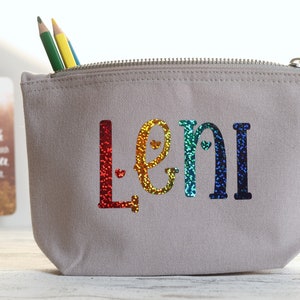 Könnte beinhalten: Eine graue Canvas-Kosmetiktasche mit Reißverschluss, auf der der Name "Leni" in regenbogenfarbenen Glitzerbuchstaben geschrieben steht. Die Buchstaben haben kleine Herzen.