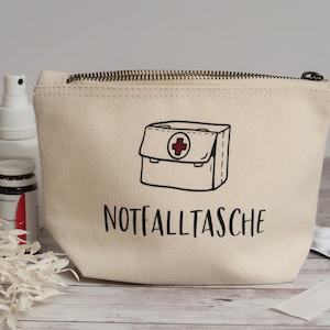 Könnte beinhalten: Eine beige Canvas-Reißverschlusstasche mit einer schwarz-weißen Illustration eines Erste-Hilfe-Kastens und dem Text "NOTFALLTASCHE".