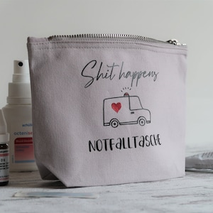 May include: A gray canvas zippered pouch with the text "Shit happens" and a cartoon ambulance with a red heart. The text "Notfalltasche" is printed below the ambulance.
