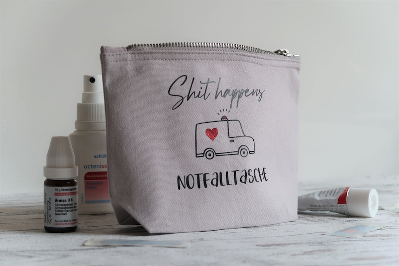 May include: A gray canvas zippered pouch with the text "Shit happens" and a cartoon ambulance with a red heart. The text "Notfalltasche" is printed below the ambulance.