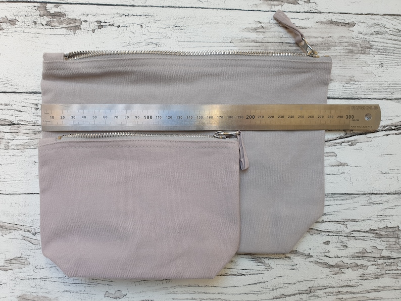 May include: Two zippered pouches made of a light gray canvas fabric. The pouches are nested inside each other, with the smaller pouch on top. A ruler is placed on top of the pouches, showing the larger pouch is approximately 30 inches wide.