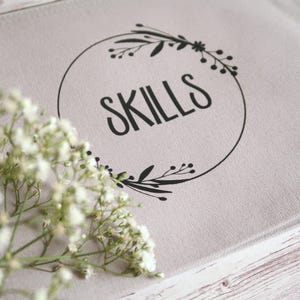 Könnte beinhalten: Eine hellgraue Canvas-Tasche mit dem Wort "SKILLS" in einem schwarzen, kreisförmigen Blumendesign. Ein Zweig weißer Blumen befindet sich im Vordergrund, vor einem hellen Holzuntergrund.