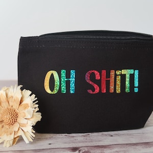 Könnte beinhalten: Schwarze Reißverschlusstasche mit dem Text "OH SHIT!" in Regenbogen-Glitzer-Schrift. Die Tasche ist rechteckig und hat einen schwarzen Reißverschluss. Eine getrocknete Blume befindet sich im Vordergrund.