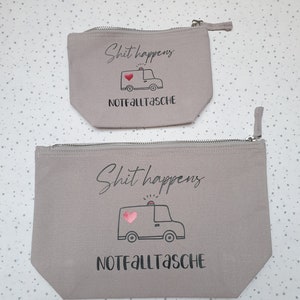 May include: Two gray canvas zippered pouches with the text "Shit happens" and a cartoon ambulance with a pink heart. The text "Notfalltasche" is printed below the ambulance.