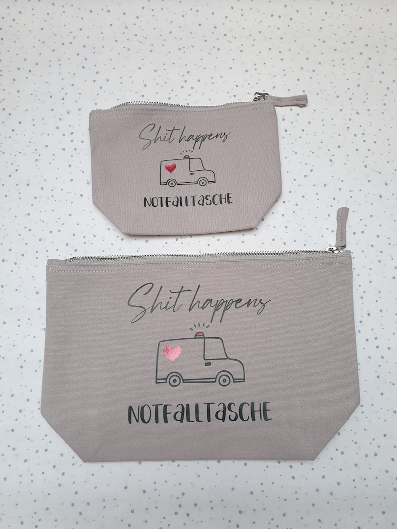May include: Two gray canvas zippered pouches with the text "Shit happens" and a cartoon ambulance with a pink heart. The text "Notfalltasche" is printed below the ambulance.