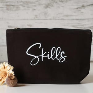 Könnte beinhalten: Schwarze Canvas-Kosmetiktasche mit dem Wort "Skills" in weißer, kursiv geschriebener Schrift.