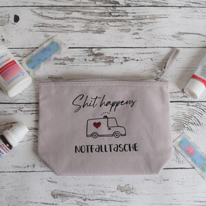 May include: A light gray canvas zippered pouch with the text "Shit happens" and a cartoon ambulance with a red heart. The pouch also has the text "Notfalltasche" printed on it. The pouch is surrounded by various first aid items, including a spray bottle, a tube of cream, and bandages.