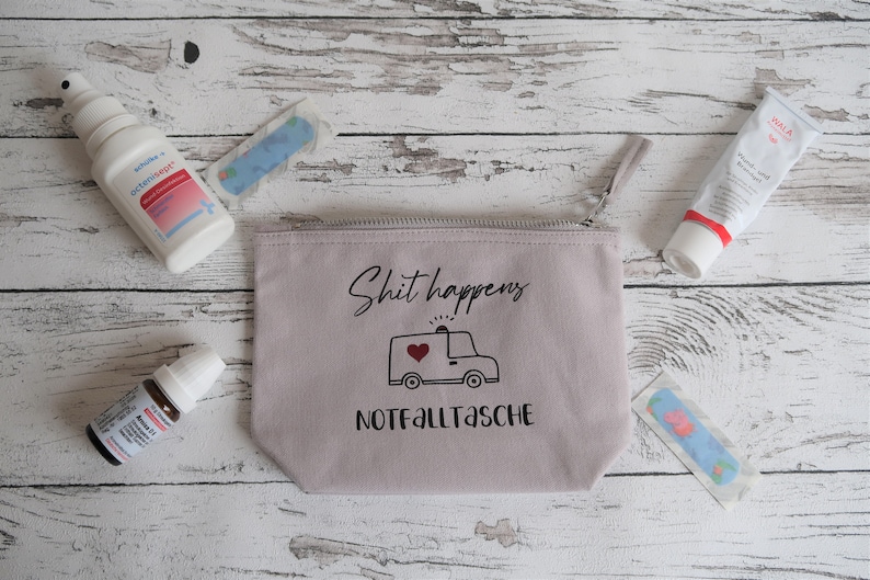 May include: A light gray canvas zippered pouch with the text "Shit happens" and a cartoon ambulance with a red heart. The pouch also has the text "Notfalltasche" printed on it. The pouch is surrounded by various first aid items, including a spray bottle, a tube of cream, and bandages.