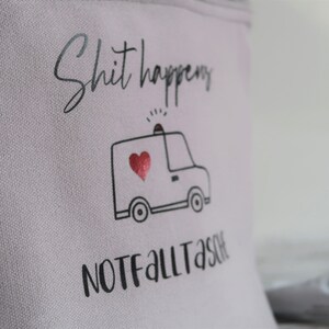 May include: A light gray canvas pouch with the text "Shit happens" and a black and white illustration of an ambulance with a red heart. The text "NOT FALL TASTE" is printed below the ambulance.