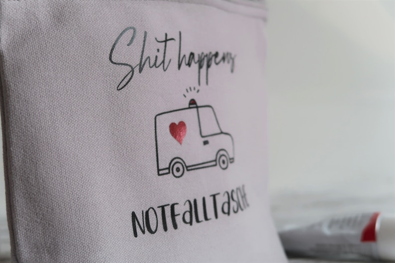 May include: A light gray canvas pouch with the text "Shit happens" and a black and white illustration of an ambulance with a red heart. The text "NOT FALL TASTE" is printed below the ambulance.