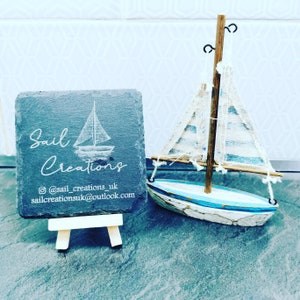 May include: A black slate coaster with the text "Sail Creations" and a sailboat graphic. The coaster is on a small wooden easel. The text "@sail_creations_uk" and "sailcreationsuk@outlook.com" is also on the coaster.