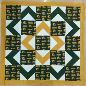 May include: A handmade quilt featuring a repeating pattern of green and gold squares. The squares display the word "PACKERS" and a football helmet. The quilt has a yellow border and is likely a sports-themed item.