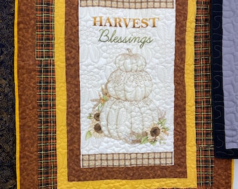 May include: A quilted wall hanging with a harvest theme. The central panel features a stacked pumpkin design with the words "HARVEST Blessings". The quilt has a white background with brown, yellow, and plaid borders.