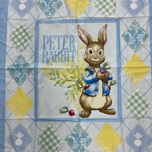 May include: A rectangular fabric cloth featuring a central illustration of Peter Rabbit wearing a blue jacket and holding a radish. The text Peter Rabbit is printed in blue. The border consists of a diamond grid pattern with yellow, green gingham, and blue floral squares.