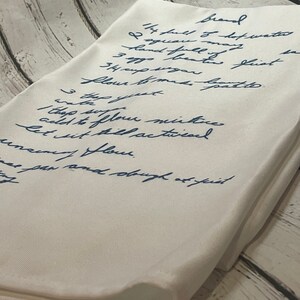 May include: A handwritten recipe for bread on a white linen napkin. The recipe includes ingredients like hot water, flour, sugar, and yeast. The recipe is written in blue ink.