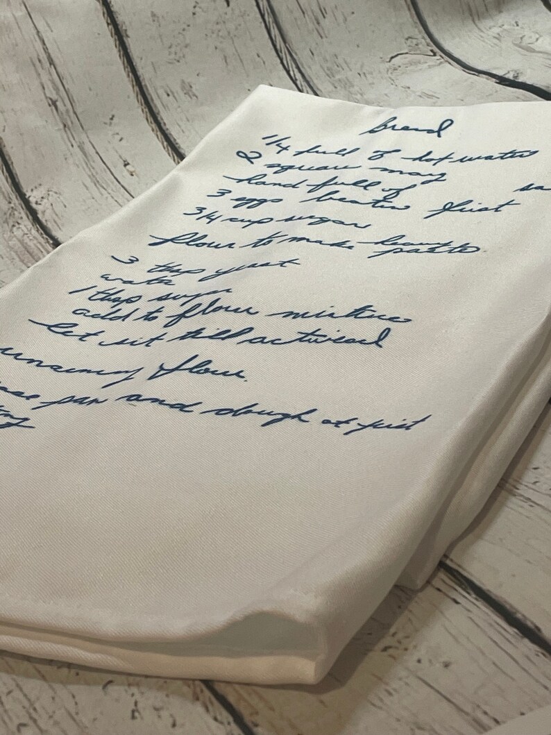 May include: A handwritten recipe for bread on a white linen napkin. The recipe includes ingredients like hot water, flour, sugar, and yeast. The recipe is written in blue ink.