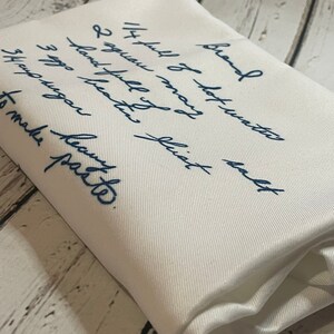 May include: A white fabric with blue ink handwritten instructions for making a recipe. The text reads "Brand Soy full of hot water 2 aguan beaten fist hand full of 3 egg sugar 5th aguan to make party."