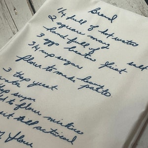 May include: A handwritten recipe for bread on a white linen tea towel. The recipe includes ingredients like hot water, flour, sugar, yeast, and eggs.