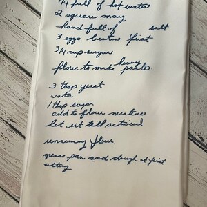 May include: A handwritten bread recipe on a white linen tea towel. The recipe includes ingredients such as hot water, shortening, salt, eggs, sugar, flour, and yeast. The instructions include mixing the ingredients, allowing the dough to rise, and greasing the pan.