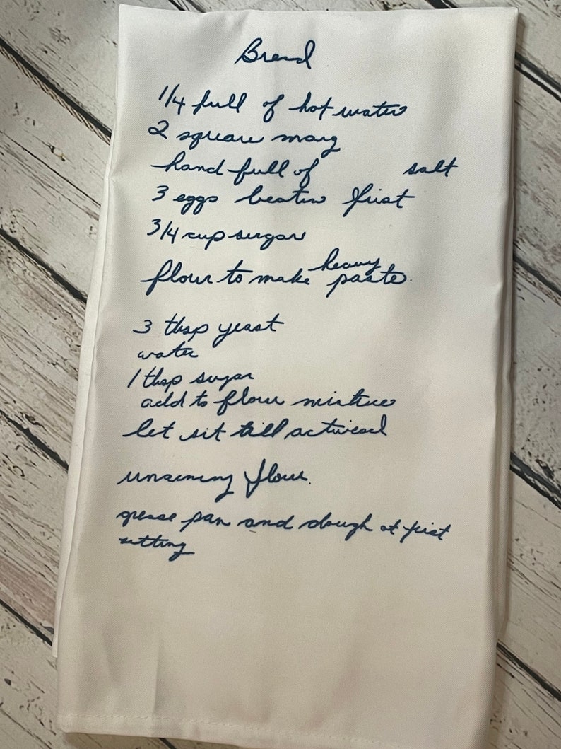 May include: A handwritten bread recipe on a white linen tea towel. The recipe includes ingredients such as hot water, shortening, salt, eggs, sugar, flour, and yeast. The instructions include mixing the ingredients, allowing the dough to rise, and greasing the pan.