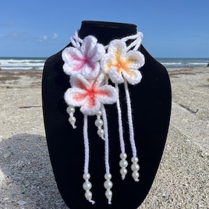 May include: A handmade crochet necklace featuring three white plumeria flowers with pink and orange accents. The flowers are attached to a white cord, with dangling strands of white yarn and faux pearls. The necklace is displayed on a black mannequin.