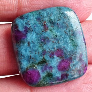 Puede incluir: Una piedra preciosa cabujón pulida de forma cuadrada que presenta un color base azul cerceta vibrante con inclusiones de color magenta intenso y púrpura incrustadas en toda la superficie, con un acabado liso y brillante y esquinas redondeadas.