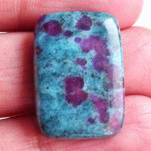 Puede incluir: Una piedra preciosa cabujón rectangular con una superficie pulida, un color base azul cerceta vibrante e inclusiones irregulares de color magenta intenso dispersas por toda la piedra, creando un aspecto moteado distintivo.