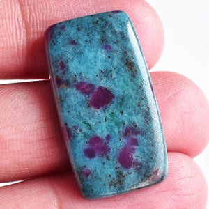 Puede incluir: Una piedra preciosa pulida de forma rectangular con un color base azul verdoso vibrante y motas opacas de color magenta intenso y púrpura distribuidas por toda la superficie. La piedra presenta un acabado liso y brillante con esquinas redondeadas.
