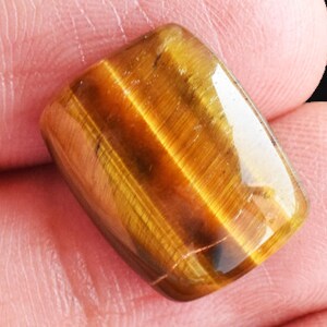 May include: A polished, rectangular tiger's eye gemstone featuring a chatoyant surface with alternating bands of golden-brown and dark brown colors, displaying a smooth, reflective finish and rounded edges on a flat, cabochon-style surface.
