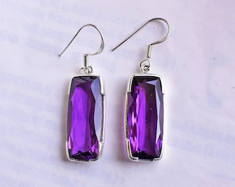 May include: A pair of rectangular purple gemstone earrings with a silver-colored metal frame and hook. The earrings have a deep purple hue and are displayed against a white background. The gemstones are cut in a rectangular shape.